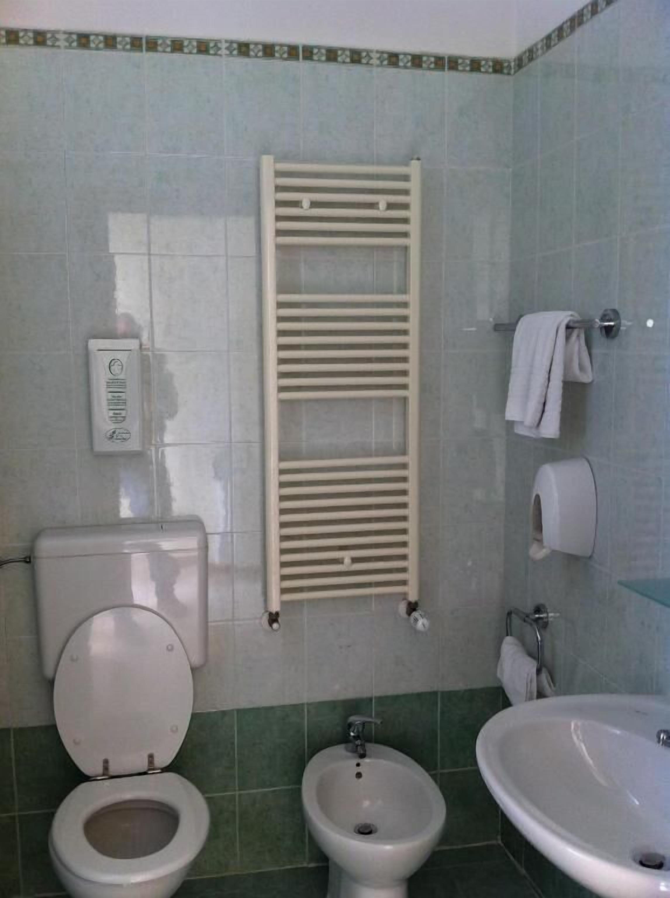 A bathroom with a toilet, bidet, sink, towel rack, and a wall-mounted toilet paper dispenser.