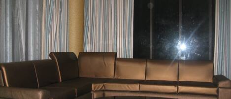 A modern living room with a brown sectional sofa, a white coffee table, and a large window with curtains.