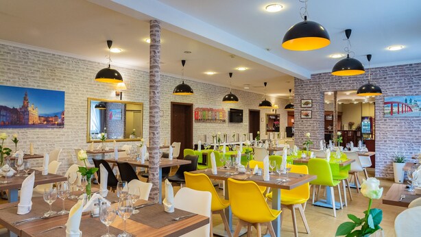 A restaurant with multiple tables, some with yellow and white chairs, and others with green and white chairs. There are black pendant lights hanging from the ceiling. - Hotel Topaz Poznań Centrum (Poznan)