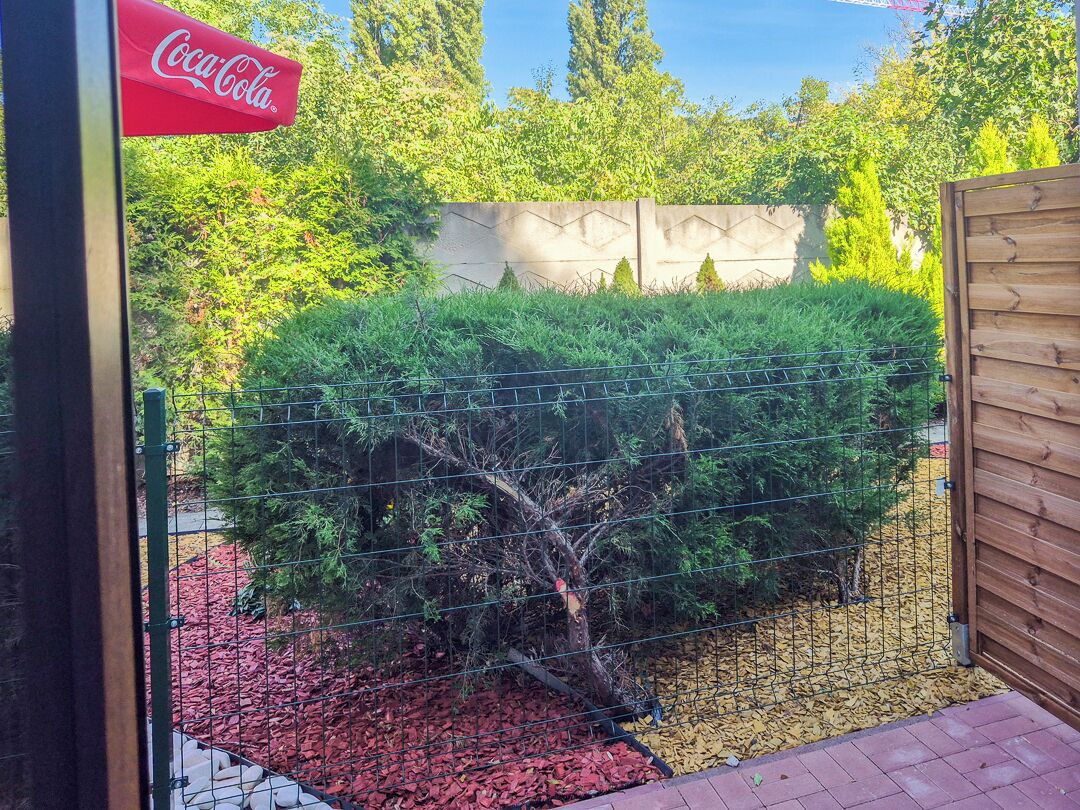 Een bord met Coca-Cola logo, een houten poort, een prikkeldraadhek en een aangelegde tuin met rode mulch en groene struiken.