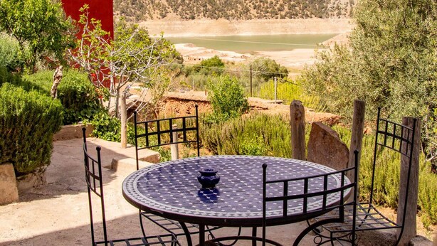 A round metal table with chairs, a blue vase, and a view of a river and mountains in the background. - Riad Diwane (Ouirgane)