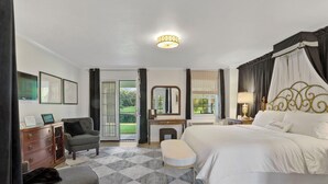 A bedroom with a large bed, a desk, a chair, and a small stool. There is a window with curtains, a mirror, and a lamp on the nightstand.