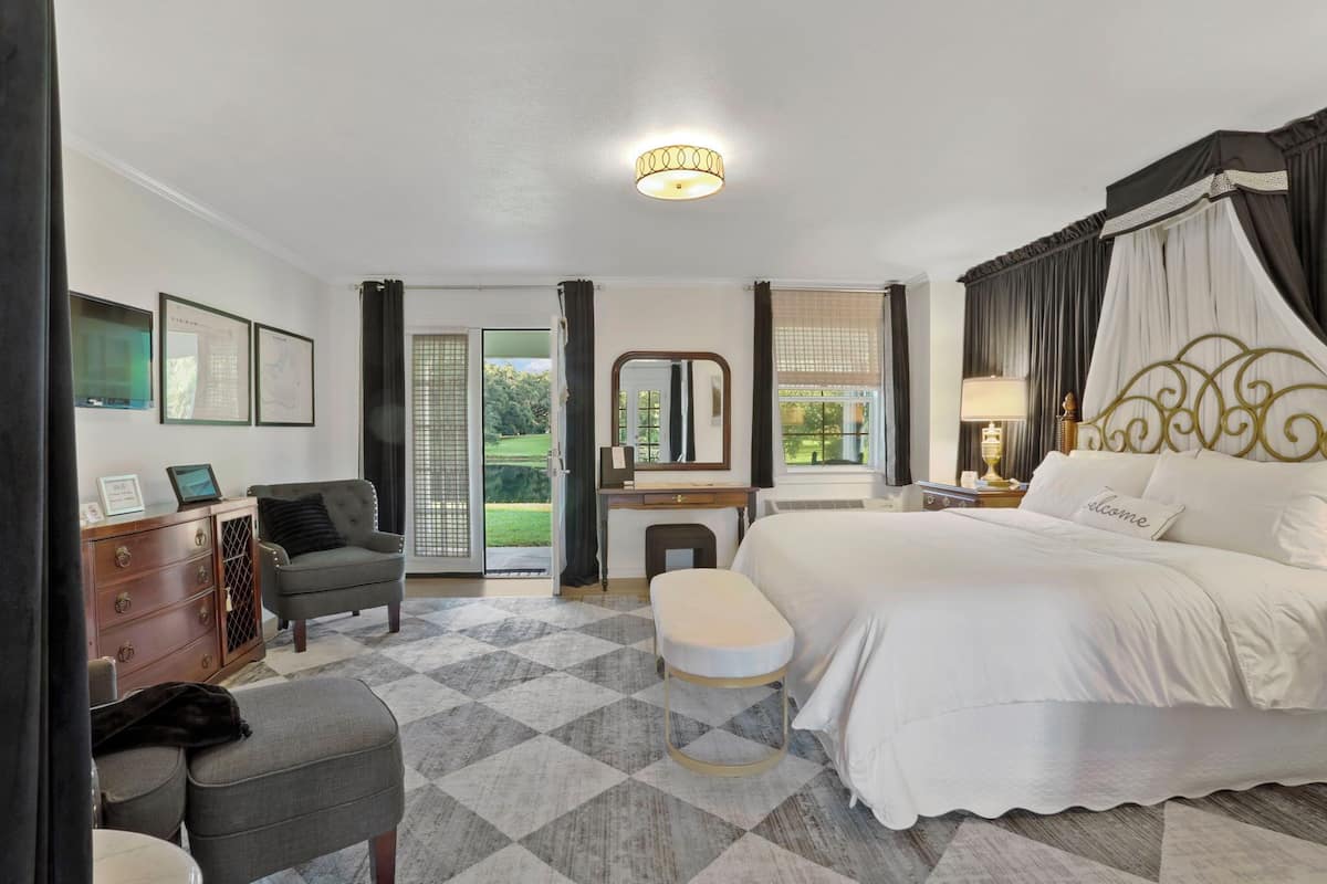 A bedroom with a large bed, a desk, a chair, and a small stool. There is a window with curtains, a mirror, and a lamp on the nightstand.
