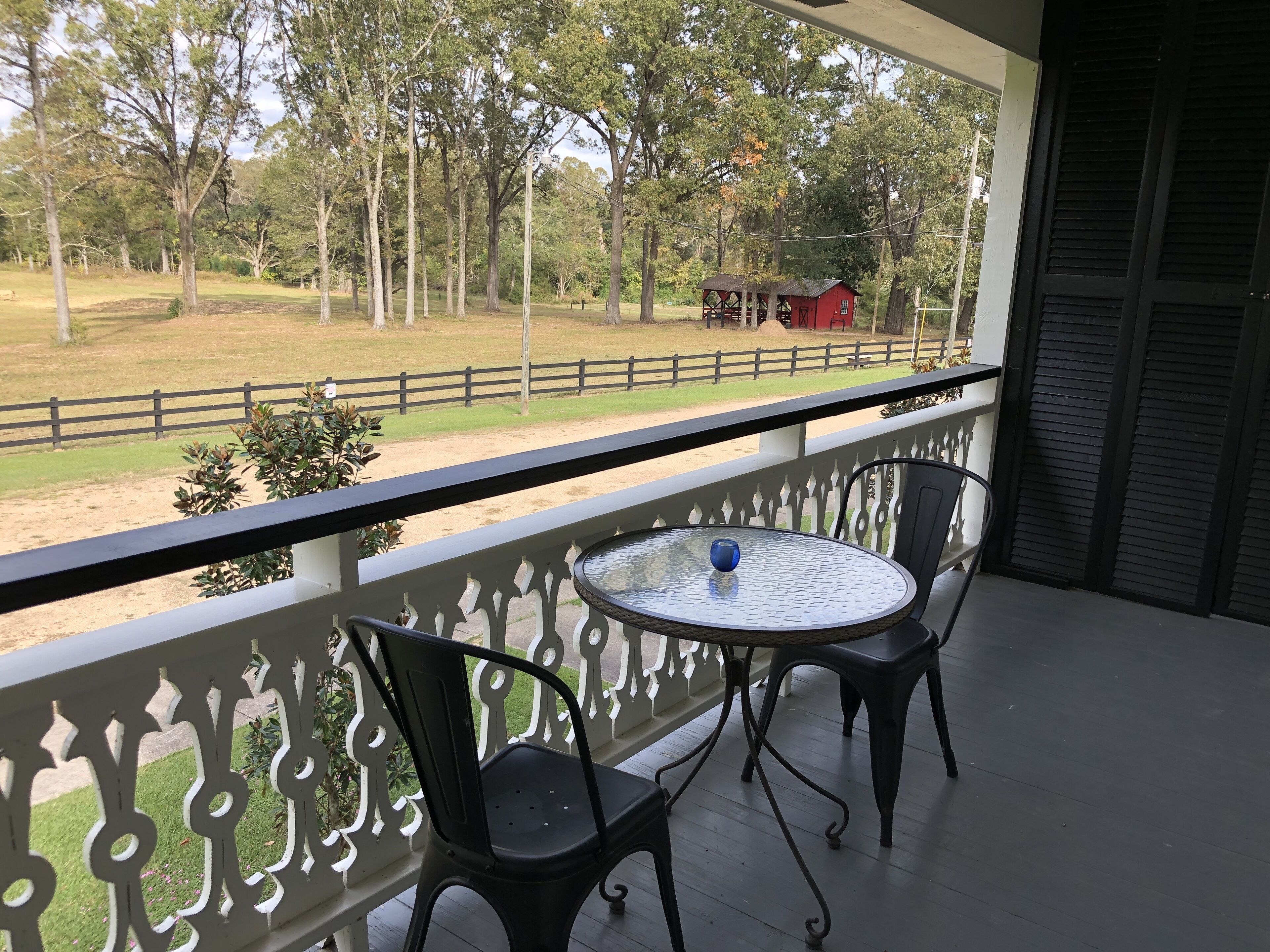 Un balcon avec une table et des chaises, donnant sur un espace clos où se trouvent des arbres et, au loin, un bâtiment rouge.