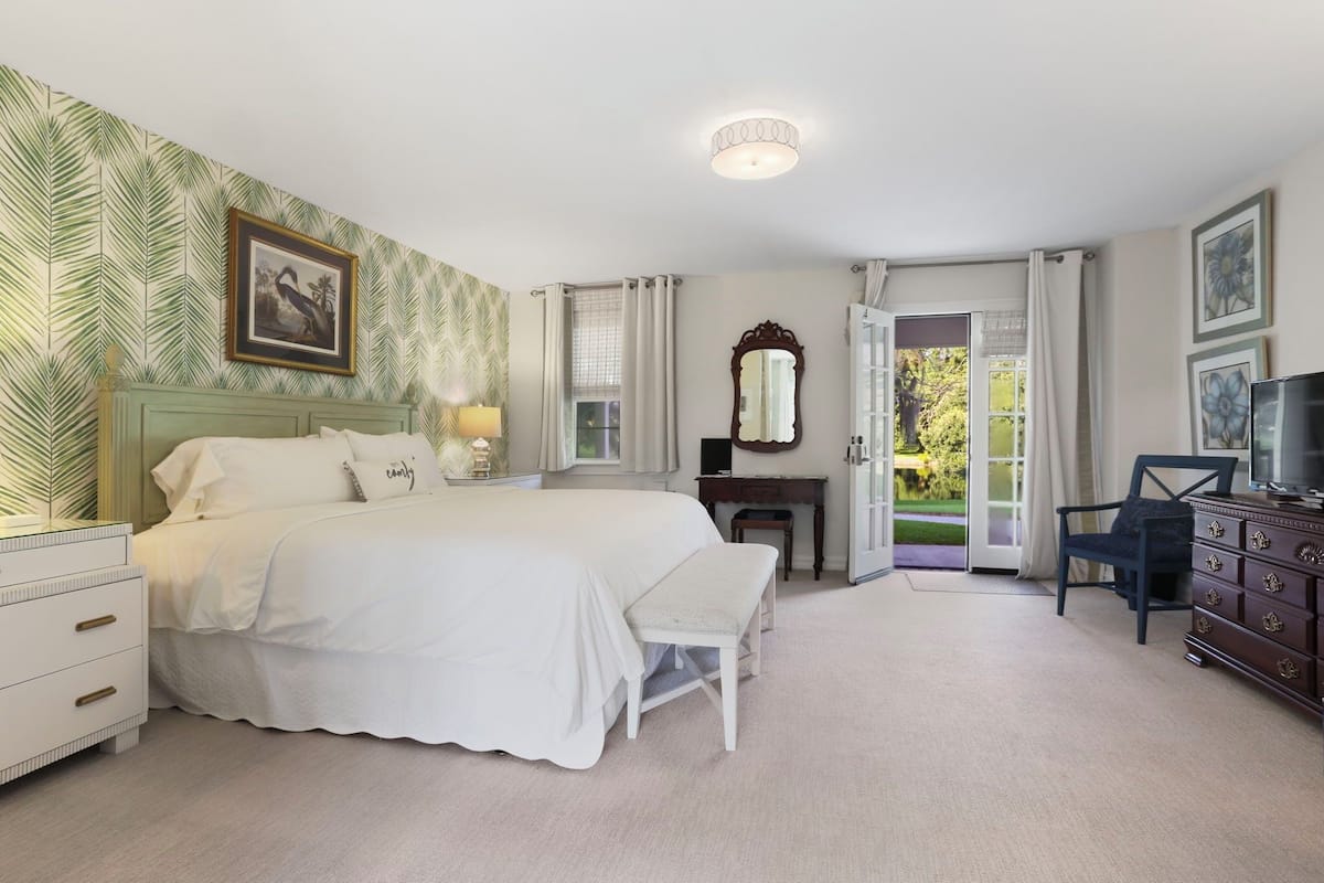 A bedroom with a large bed, a bedside table with a lamp, a dresser, a mirror, and a television.
