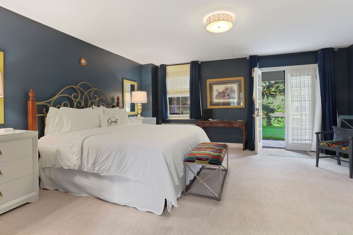 A bedroom with a large bed, bedside table, lamp, and a view of the outdoors through a window.