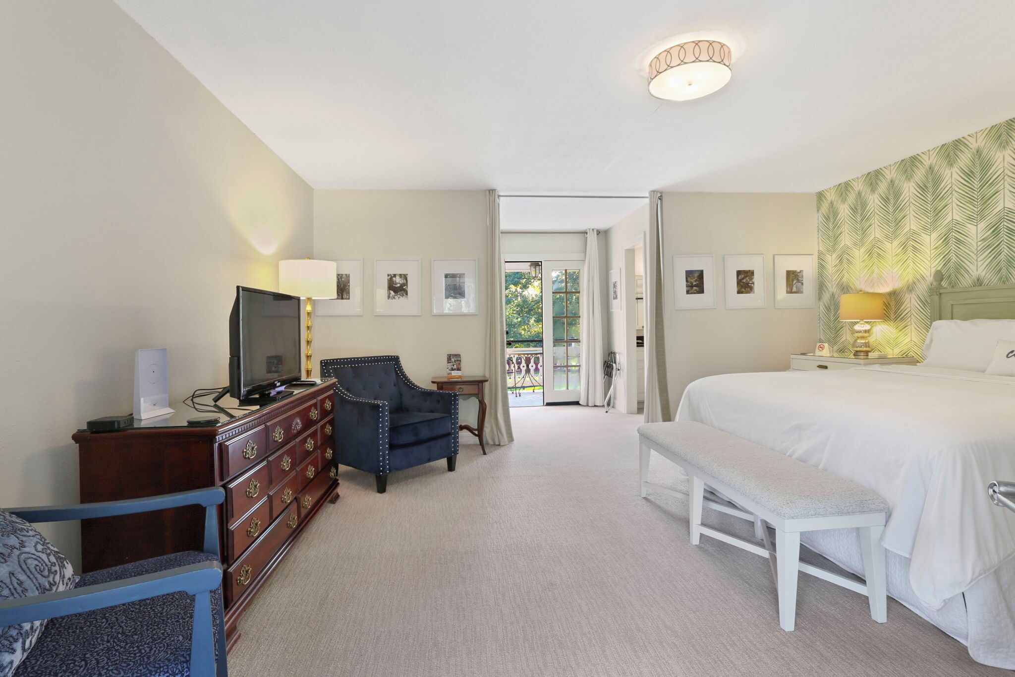 Ein Hotelzimmer mit einem großen Bett, einem Schreibtisch mit Computer, einem Sessel, einer Bank und einem Fernseher.