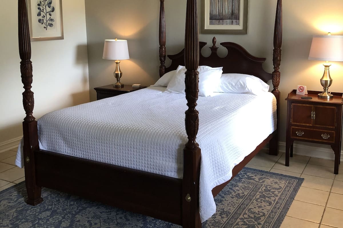 A four-poster bed with a wooden frame, a white bedspread, and a blue patterned rug. There are two bedside tables with lamps, a ceiling fan, and a framed artwork on the wall.