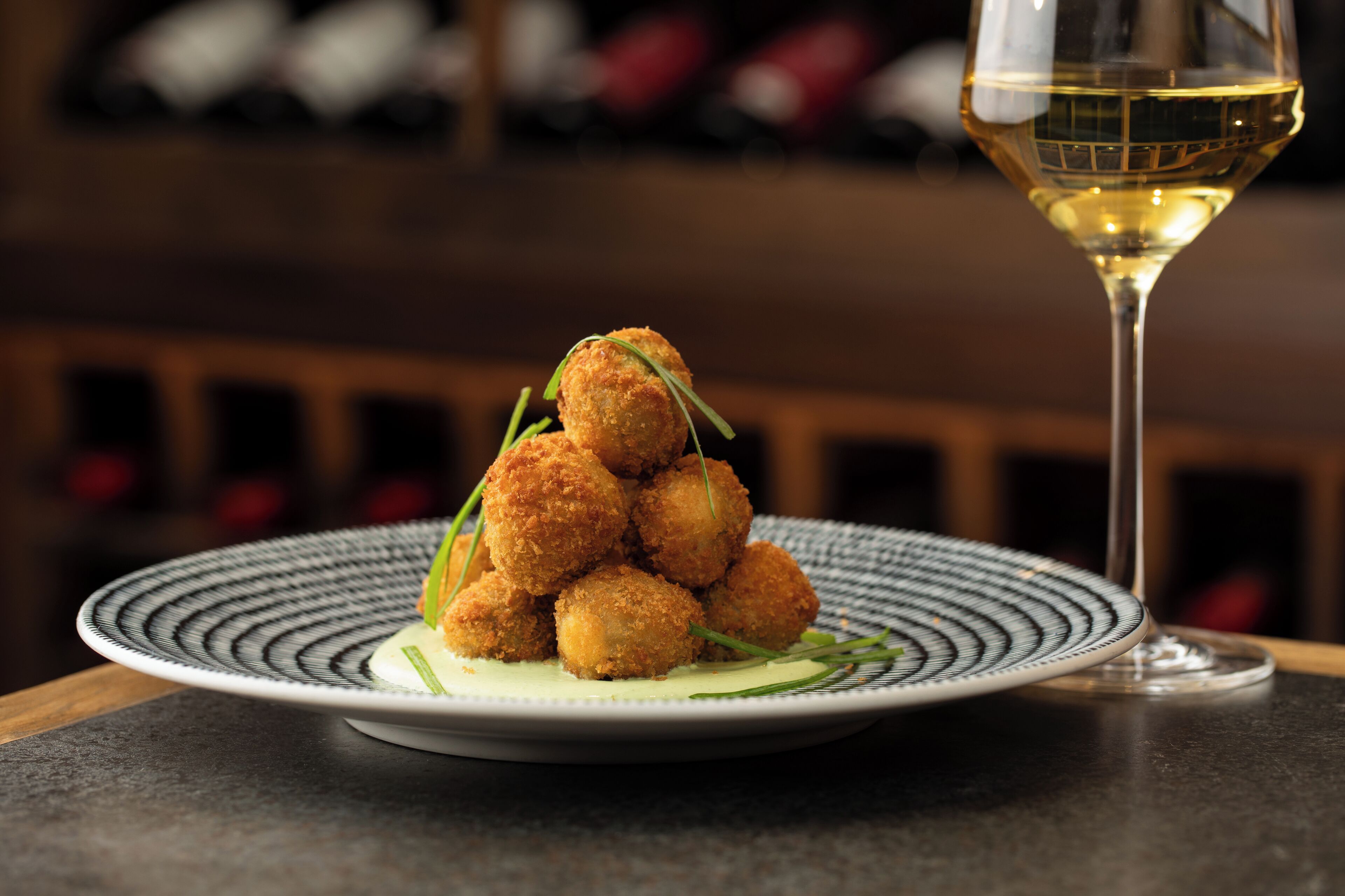 A plate of breaded and fried food items, possibly croquettes, on a patterned plate with a glass of white wine beside it.