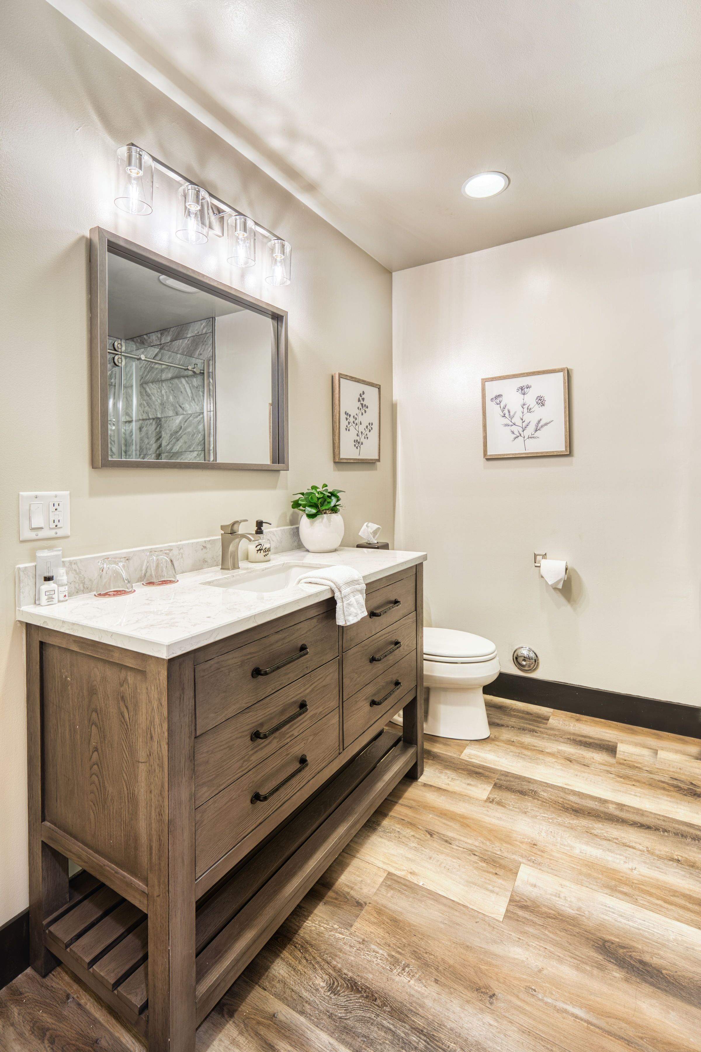 A bathroom with a wooden vanity, a large mirror, a marble countertop, a toilet, and framed botanical artwork on the wall.