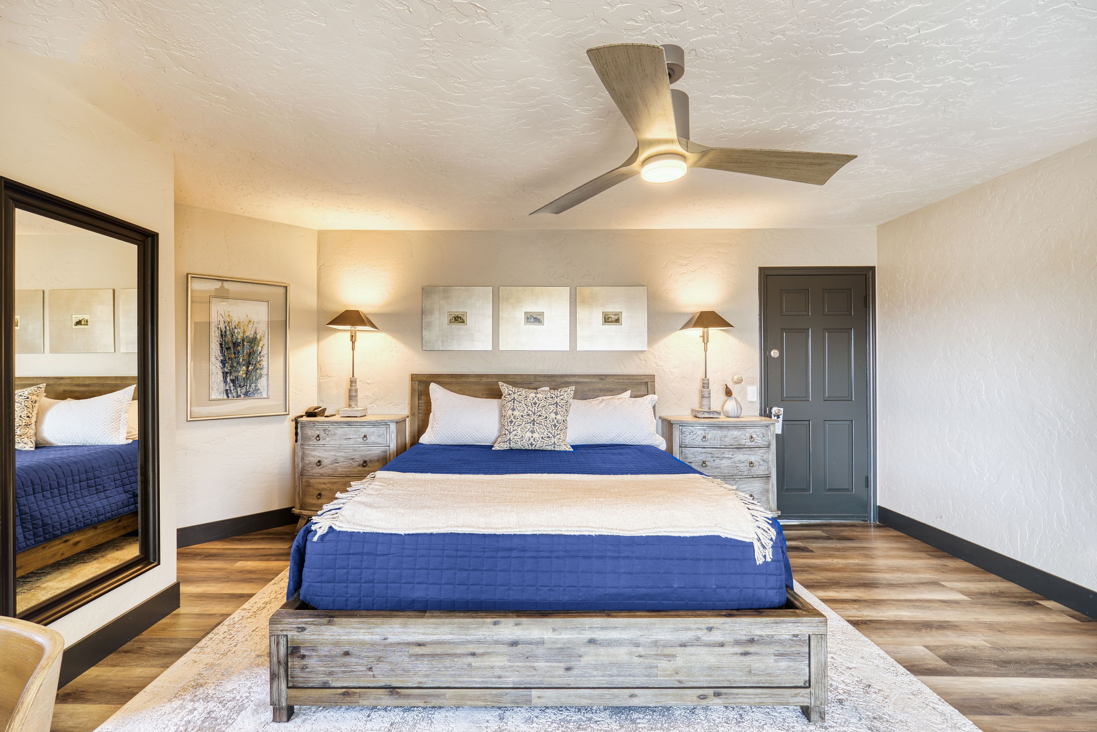 A bedroom with a large bed, a ceiling fan, bedside lamps, a door, and framed artwork on the wall.