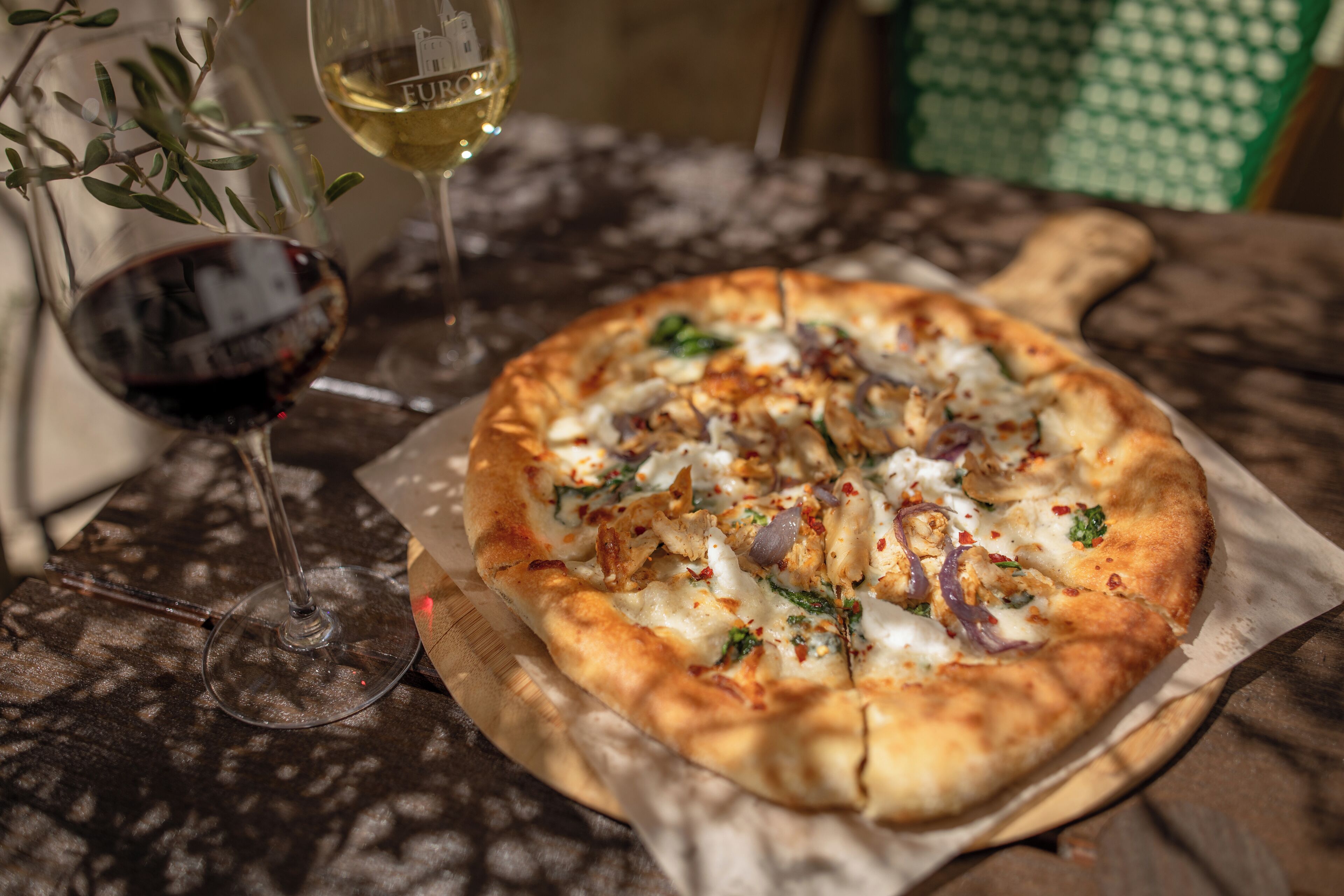 A pizza on a wooden board with a glass of red wine and a glass of white wine on the table.