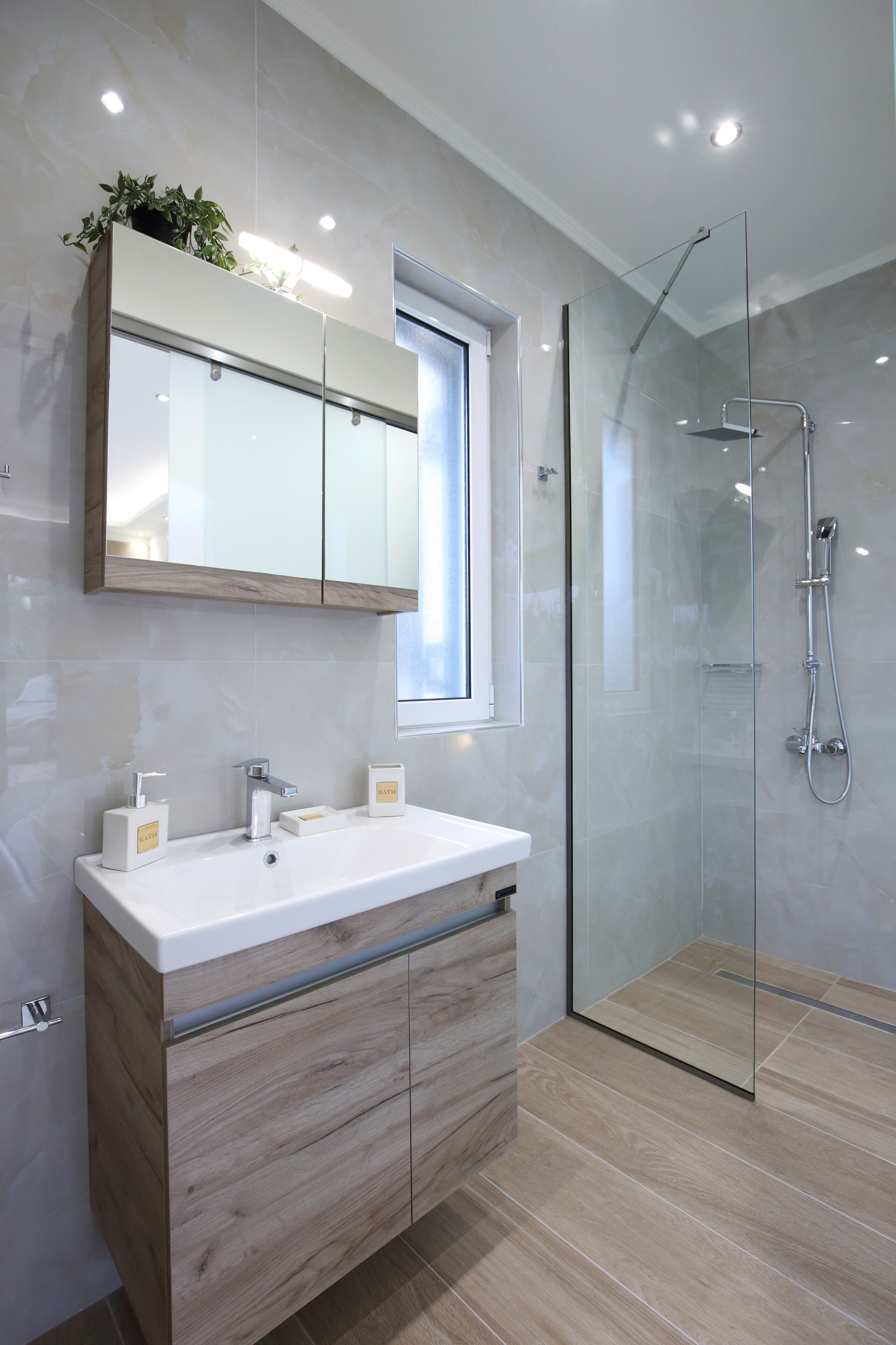 Une salle de bain moderne dotée d’une douche à parois de verre, d’un grand miroir au-dessus du lavabo et d’une fenêtre laissant entrer la lumière naturelle.