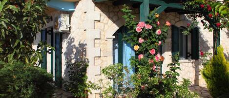 Un bâtiment en pierre avec un chemin en pierre, des volets verts et des plantes en fleurs.