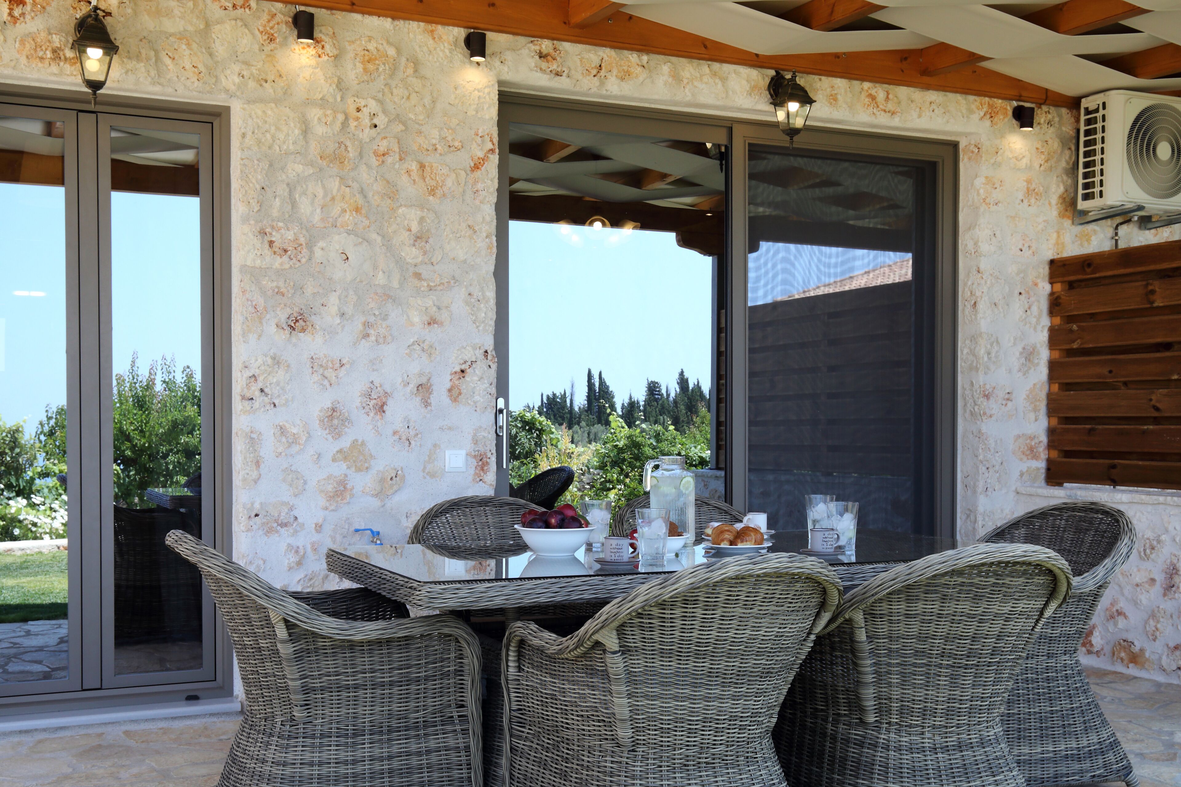 Un patio techado con mobiliario de mimbre, una mesa con jarra, cuencos y vaso, y vistas a la vegetación a través de amplios ventanales.
