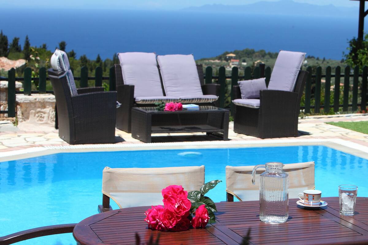 Zona junto a la piscina con mobiliario de mimbre, una mesa con jarrón y vaso, y vista al mar.