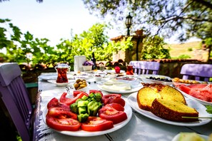 A table set outdoors with various food items including sliced tomatoes, cucumbers, and a loaf of bread. - Yasu Tenedos (Bozcaada)