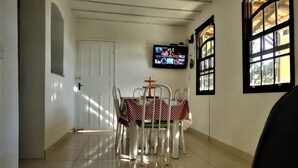 A dining area with a tiled floor, a table with chairs, a television on the wall, and a window with a view of greenery. - Vila do Sossego (Cabo Frio)