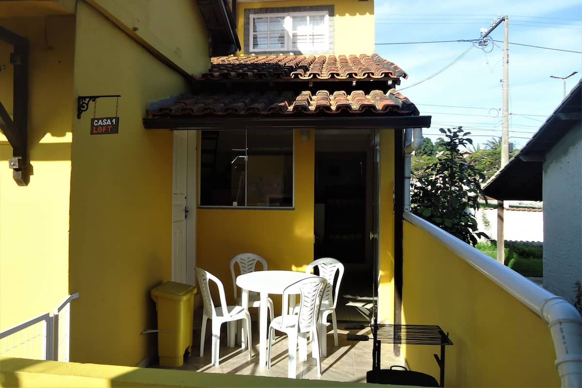 A yellow building with a tiled roof, a sign reading "CASA 1 LOFT", and a small outdoor seating area with white plastic chairs and a table.