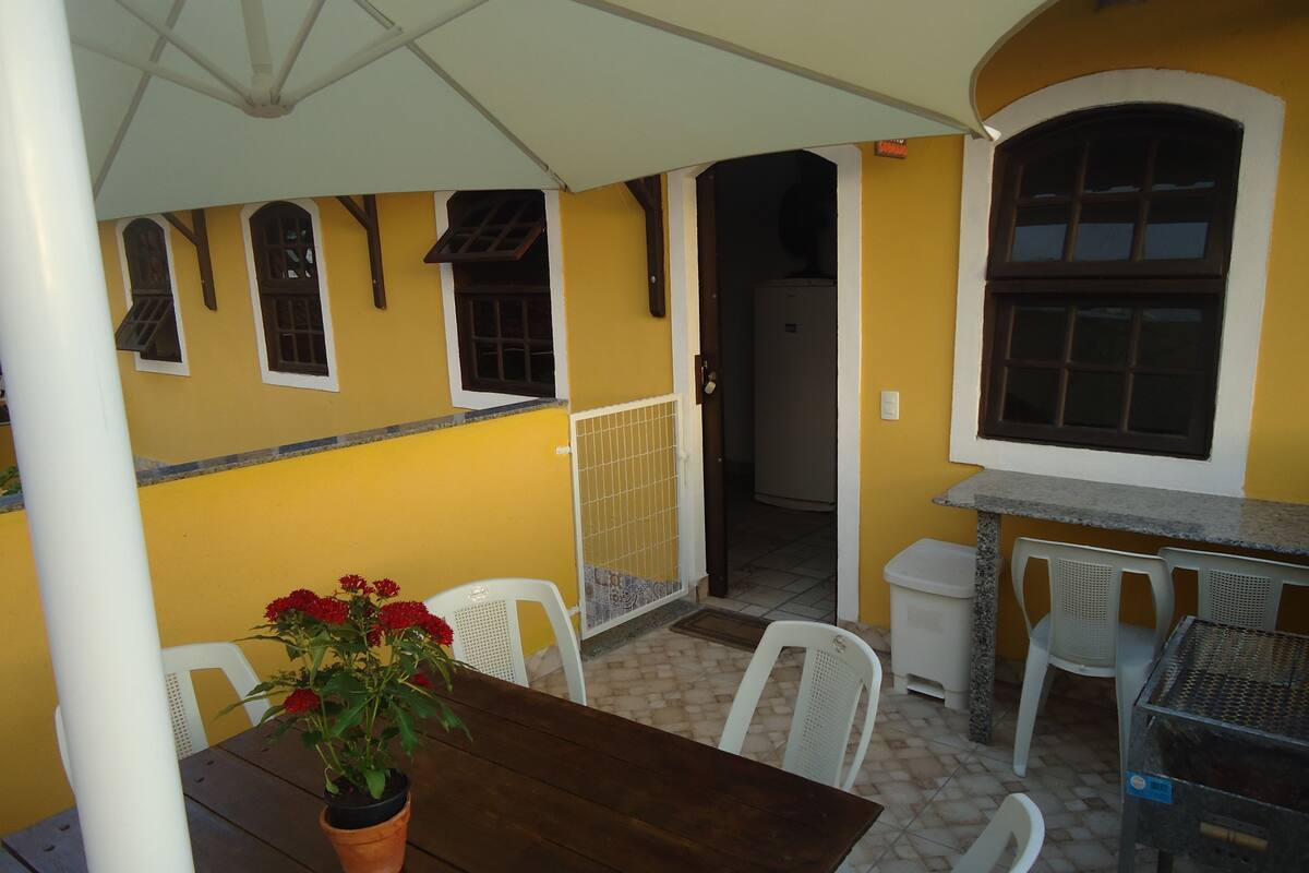 An outdoor patio area with a wooden table, white chairs, a potted plant with red flowers, and a yellow wall with windows.