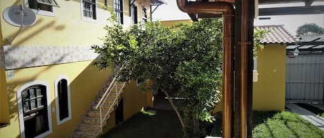 A yellow building with a staircase leading to a door, a tree in the yard, and a white wall with a window.
