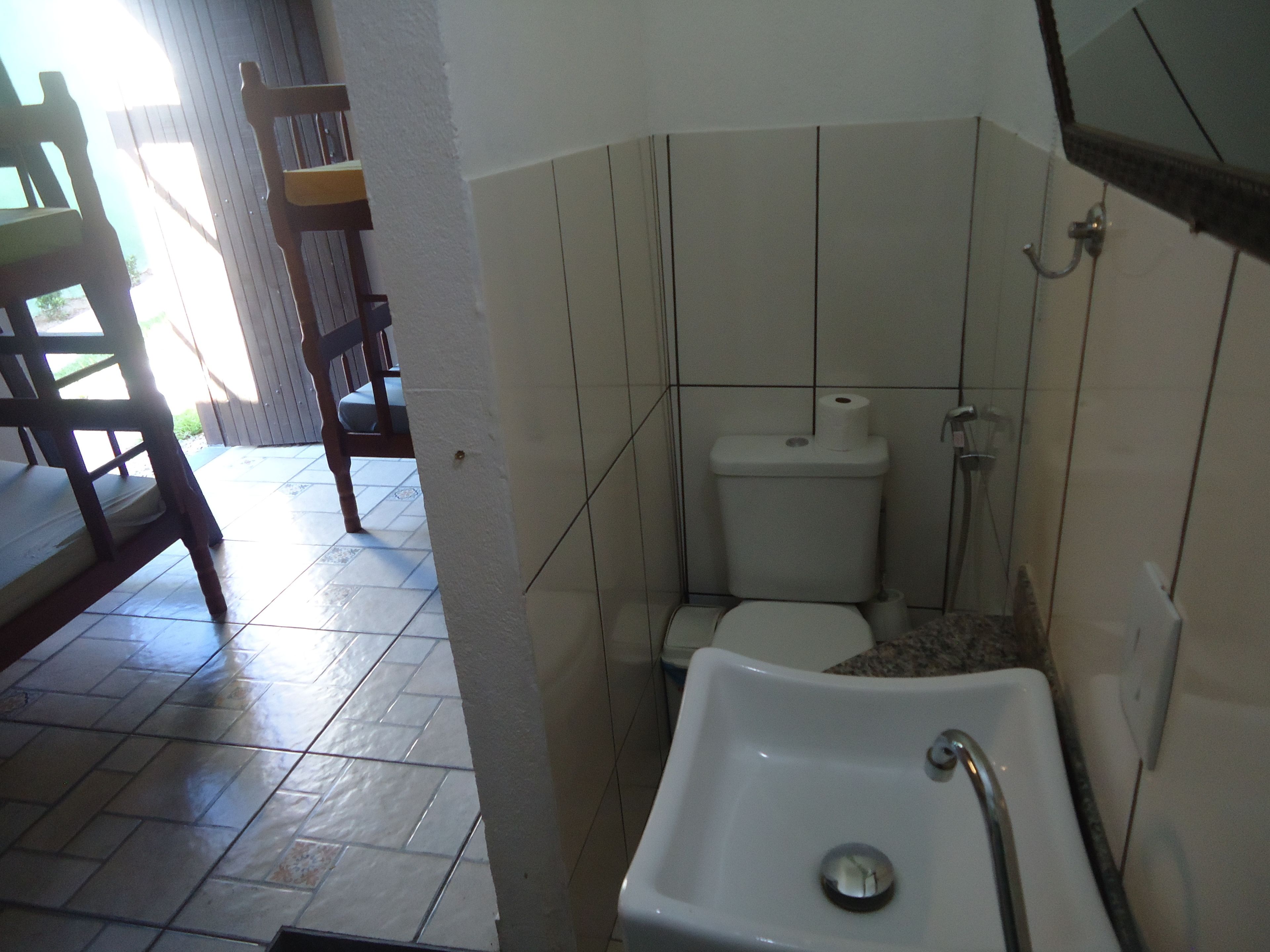 A bathroom with a toilet, sink, and shower. There is a chair outside the bathroom.