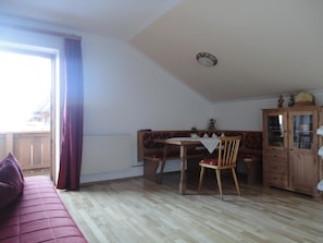 A cozy room with a wooden floor, a red sofa, a dining table with chairs, a wooden cabinet, and a window with curtains.