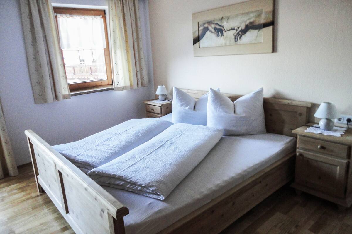Quarto com cama de madeira, roupa de cama branca, mesa de cabeceira com abajur e janela com cortinas.