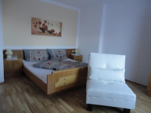 A bedroom with a wooden bed, a white armchair, a nightstand with a lamp, and a painting on the wall.