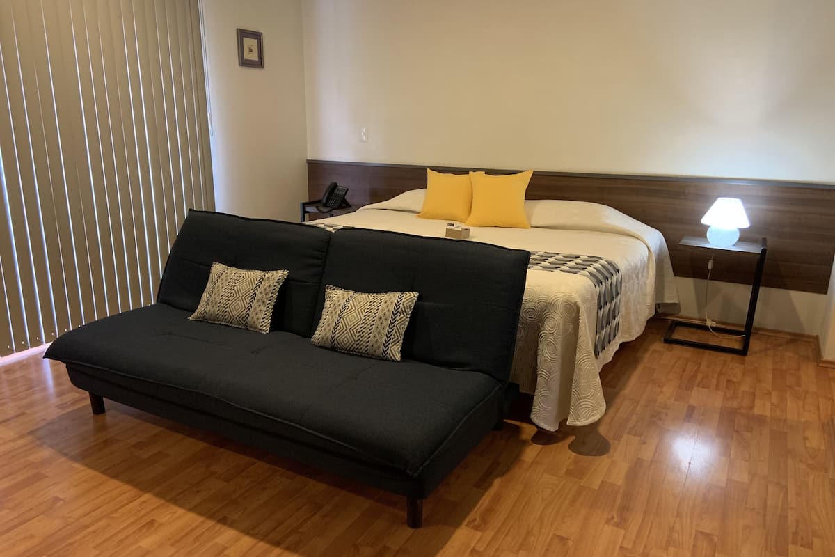 A bedroom with a bed, a black sofa, a bedside table with a lamp, and a wooden floor.