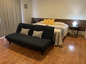 A bedroom with a bed, a black sofa, a bedside table with a lamp, and a wooden floor.