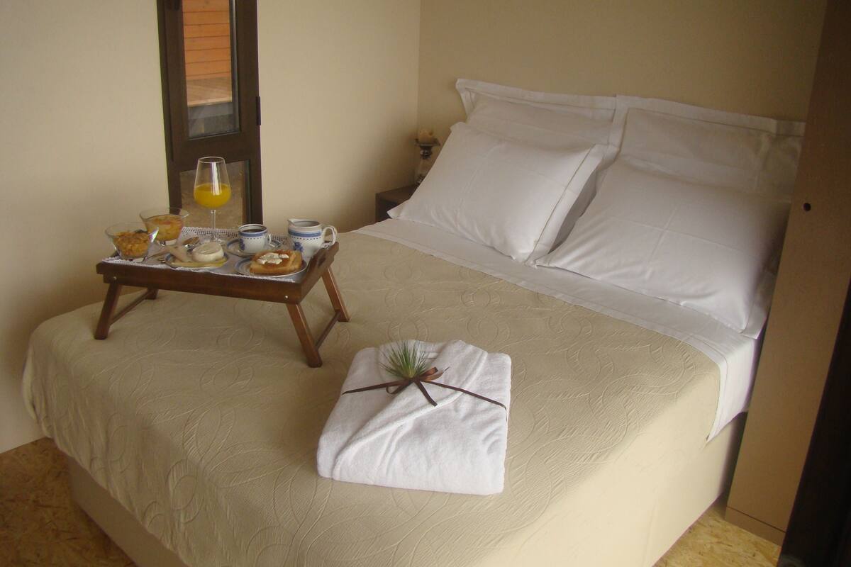 A neatly made bed with white linens, a bedside table with breakfast items, and a window with blinds.