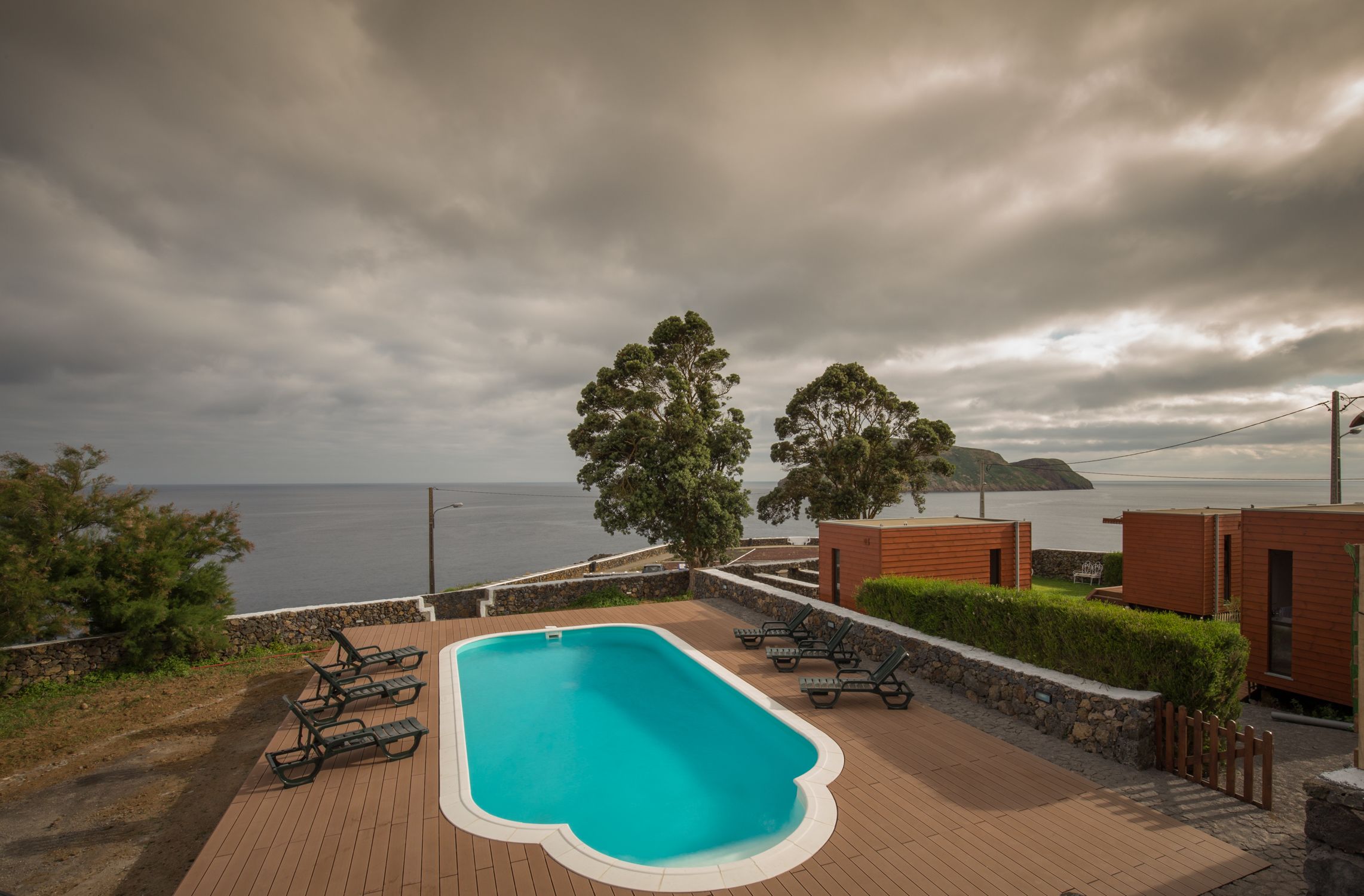 A swimming pool with lounge chairs, a stone wall, and a view of the sea.