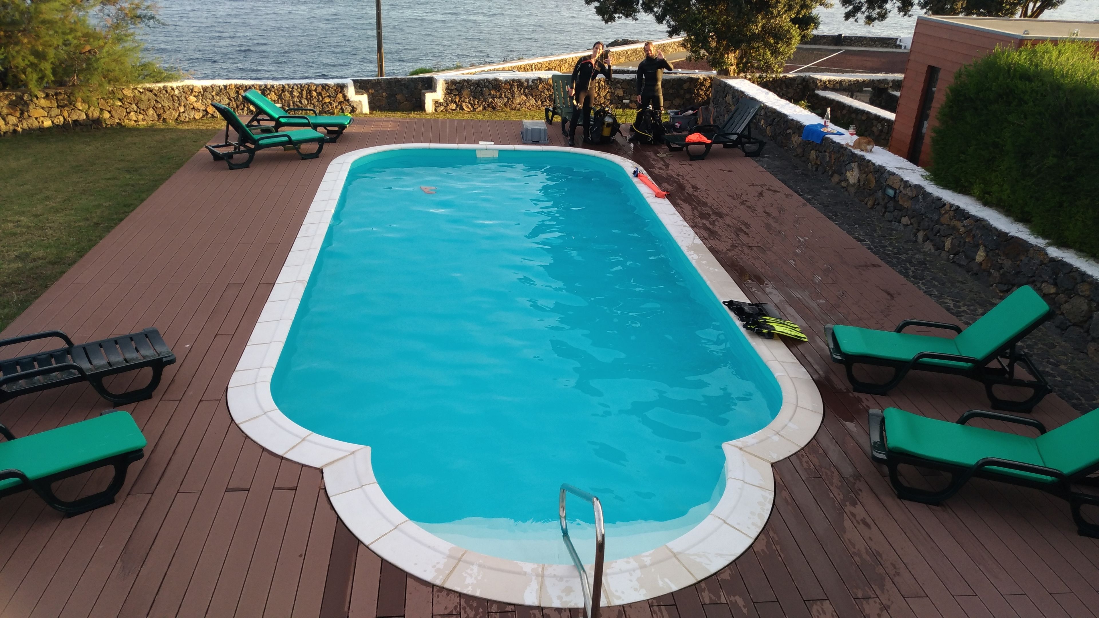 Una piscina rectangular con un deck de madera alrededor, equipada con varias sillas reclinables verdes y una pared de piedra.