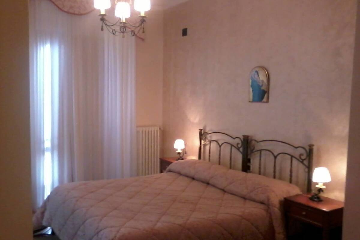 Une chambre à coucher avec un lit, deux tables de chevet avec des lampes, un radiateur, une fenêtre avec des rideaux et un tableau accroché au mur.