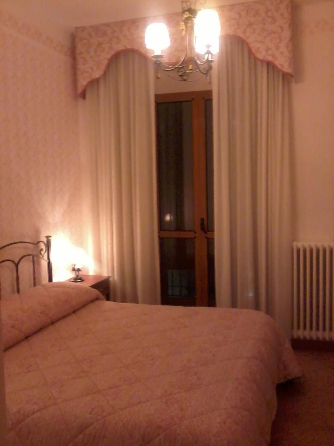 A bedroom with a bed, a nightstand, a lamp, a window with curtains, and a radiator.
