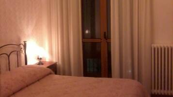 A bedroom with a bed, a nightstand, a lamp, a window with curtains, and a radiator.