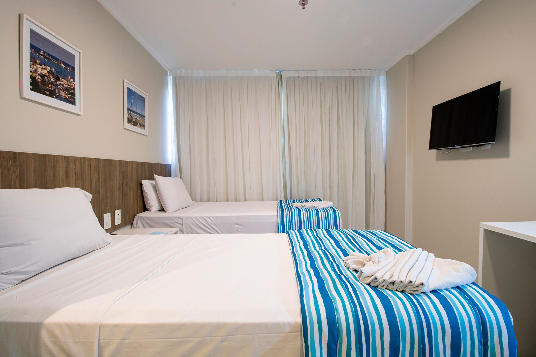 Quarto de hotel com duas camas, televisão de tela plana e uma janela grande com cortinas.