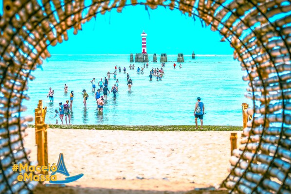 A beach scene with a lighthouse in the background, people in the water, and a hashtag #Maceió eMassa.