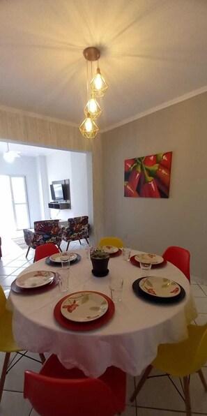 Een eethoek met een ronde tafel gedekt voor vier personen, omringd door kleurrijke stoelen. Op de muur hangt een schilderij van rode paprika's, en erboven prijkt een plafondlamp.