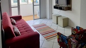 Un soggiorno con un divano rosso, una poltrona colorata, una TV a muro e una porta vetrata scorrevole che dà su un'area esterna.