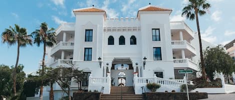 A large, white building with a symmetrical facade, multiple balconies, and a central entrance with steps.