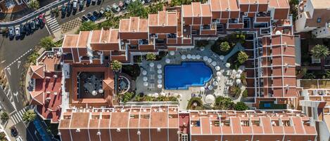 An aerial view of a hotel with a swimming pool surrounded by outdoor seating areas and multiple floors of rooms with red-tiled roofs.