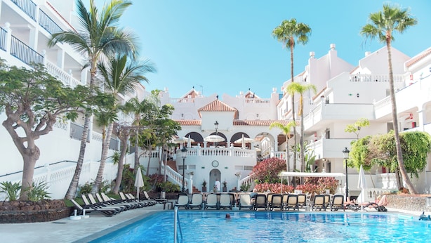Una piscina con sillas de descanso, palmeras y un edificio con balcones al fondo.