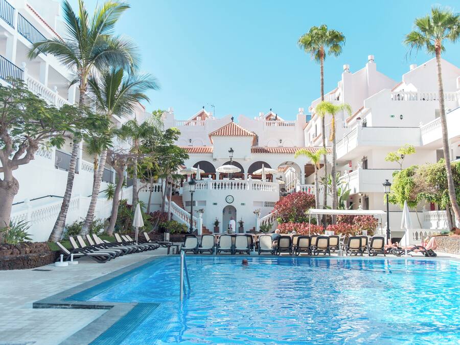 Una piscina con sillas de descanso, palmeras y un edificio con balcones al fondo.