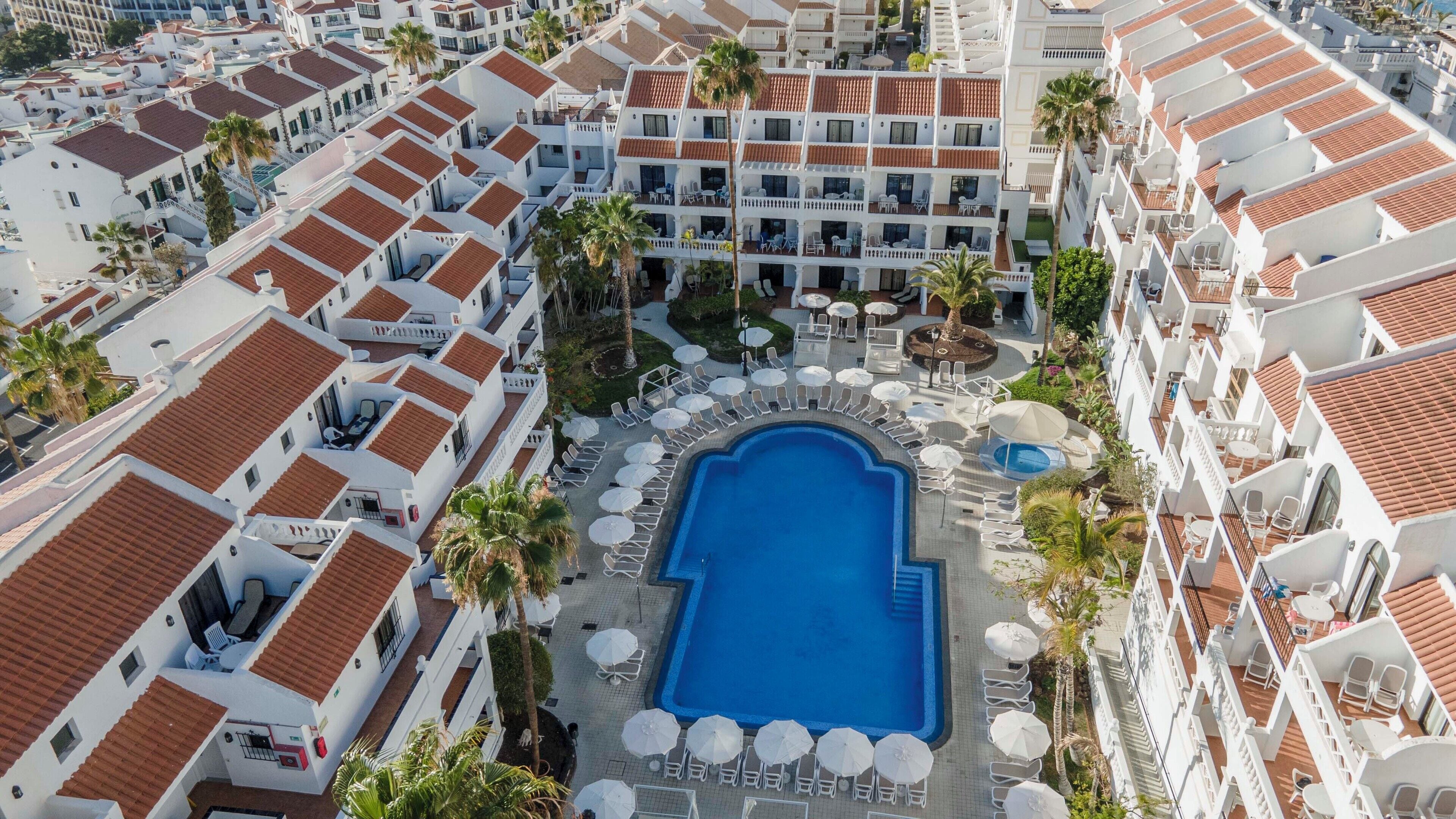 A hotel with a central swimming pool surrounded by lounge chairs and umbrellas, white buildings with red-tiled roofs, and palm trees.