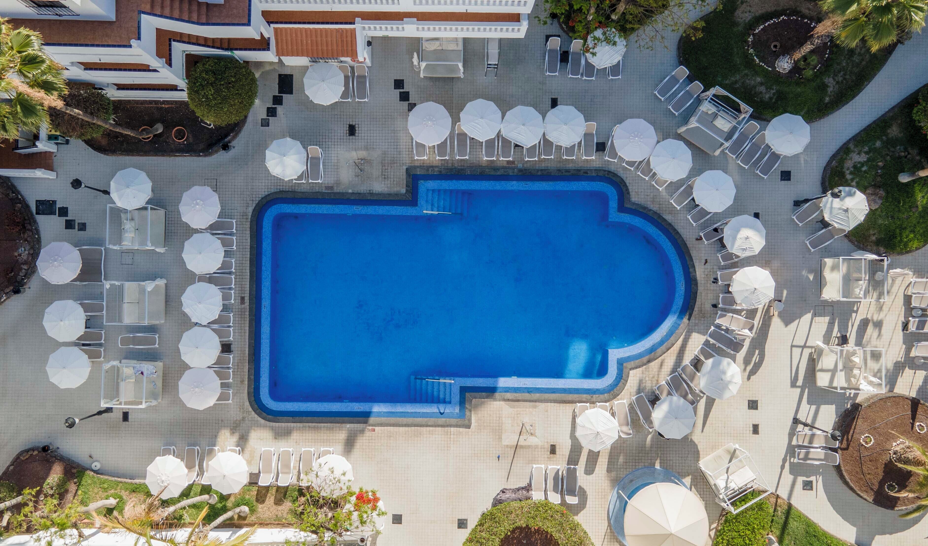 An aerial view of a swimming pool surrounded by sun loungers and umbrellas, with a tiled walkway and greenery.