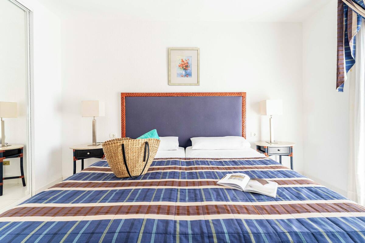 A hotel room with a large bed, two bedside tables with lamps, a woven basket, and a framed picture on the wall.