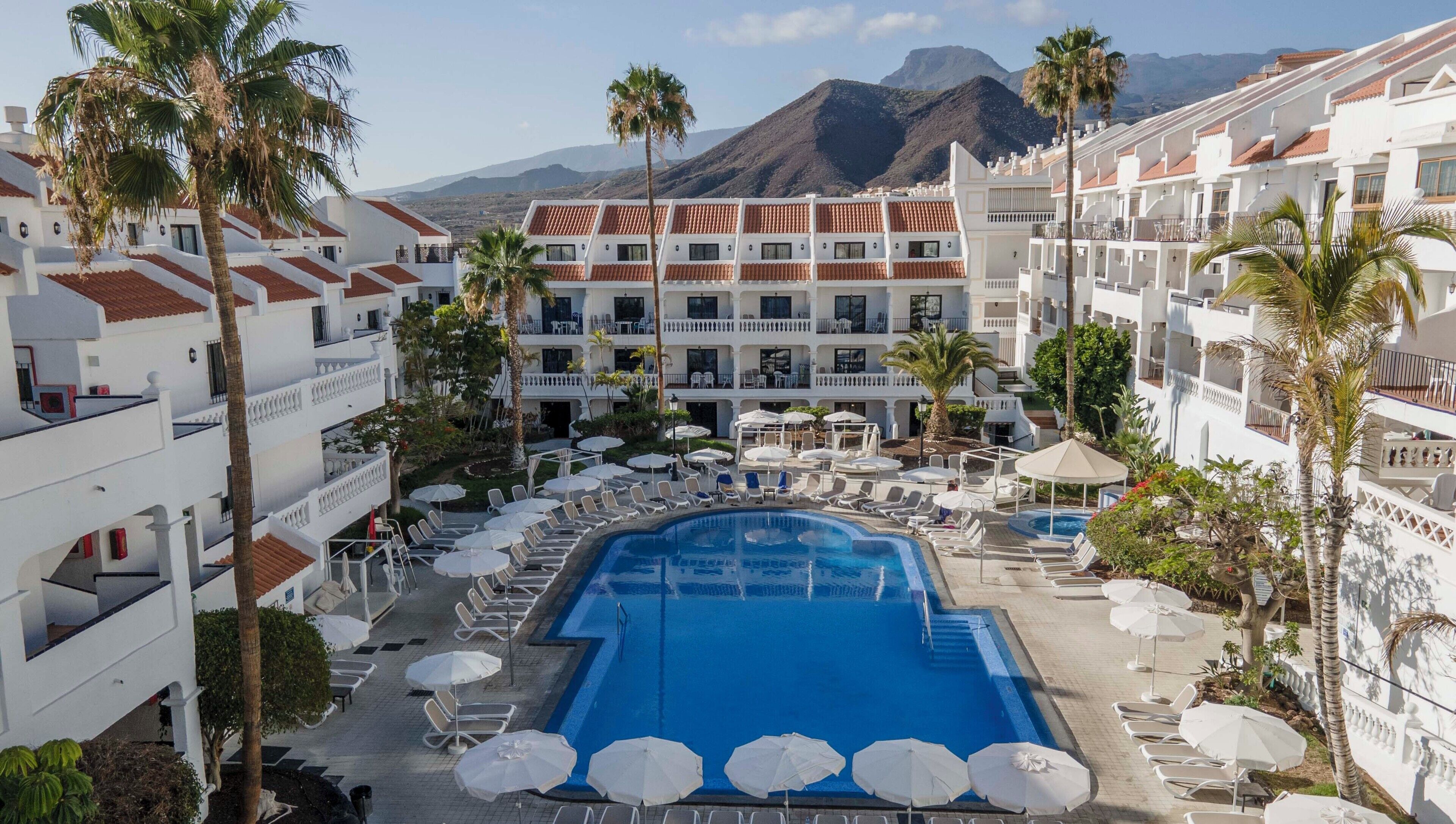 A resort with a central swimming pool surrounded by sun loungers and umbrellas, white buildings with red-tiled roofs, and palm trees.