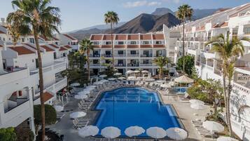 A resort with a central swimming pool surrounded by sun loungers and umbrellas, white buildings with red-tiled roofs, and palm trees.